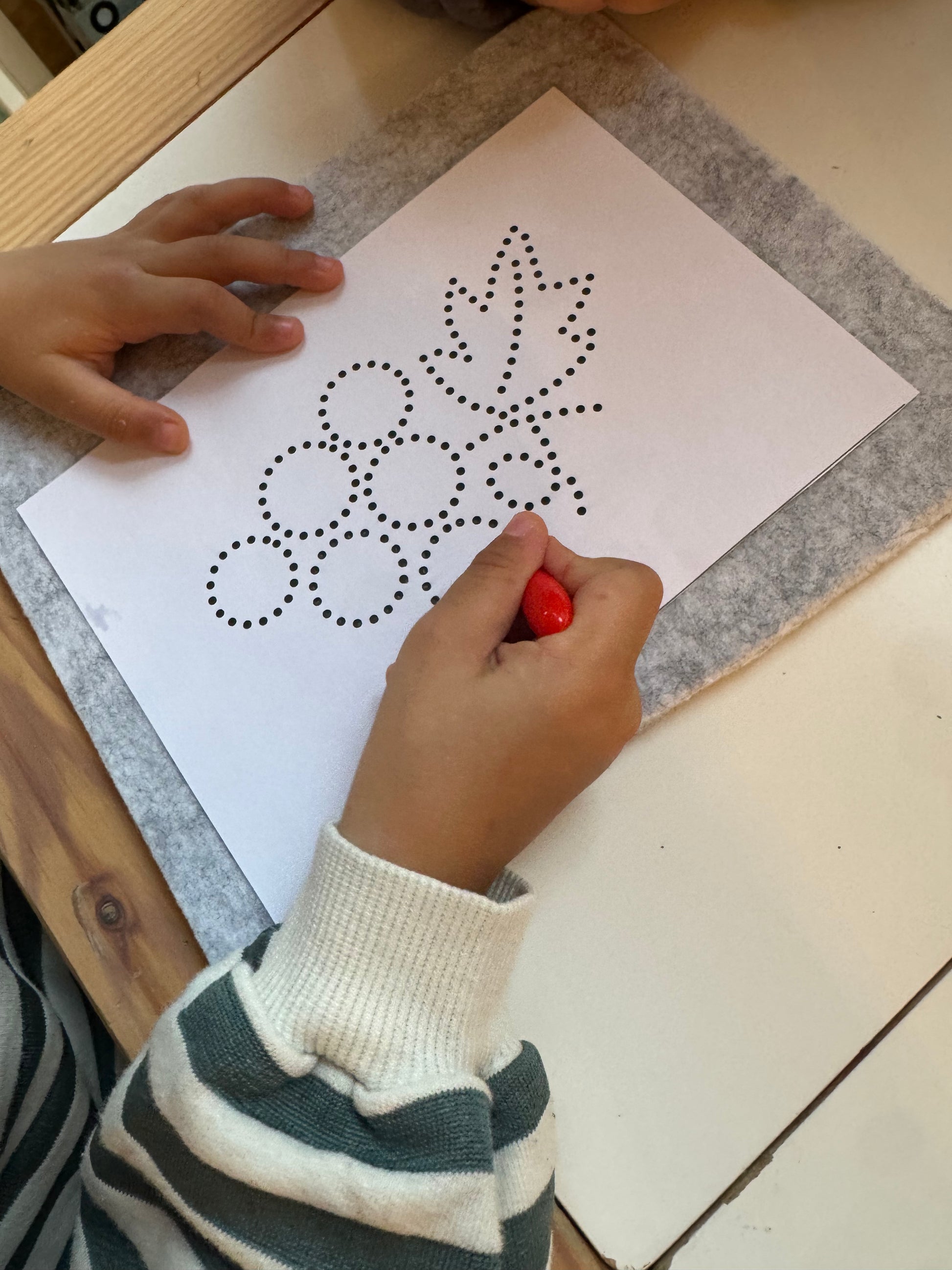 Child's hand holding a red dot marker near a paper with a dot pattern on a wooden table.