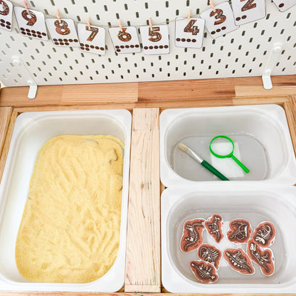 Children's play area with trays containing play dough, water, and butterfly figures, with number cards hanging above.